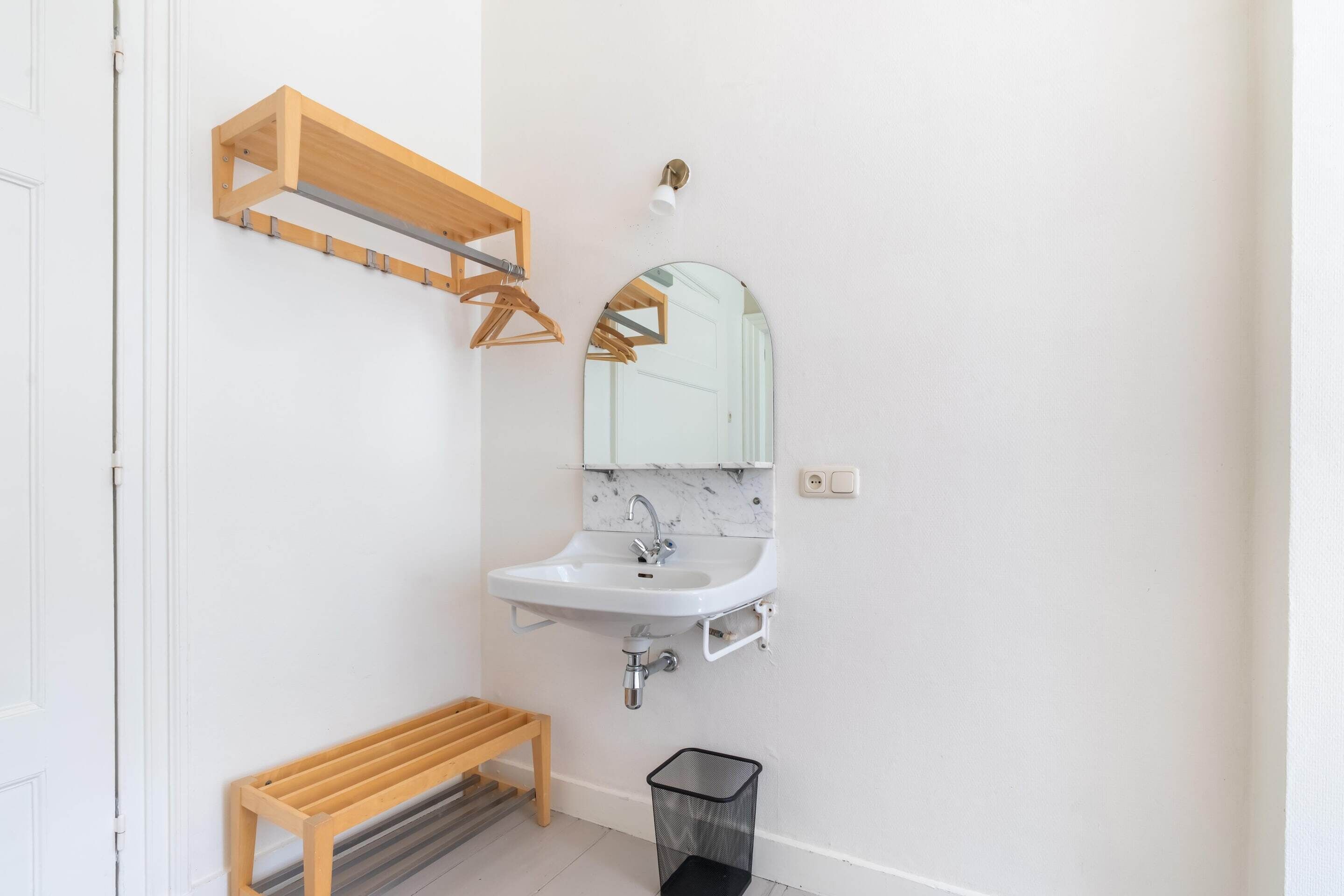 A bathroom with a white sink, a mirror, a wooden bench, and a black trash bin.