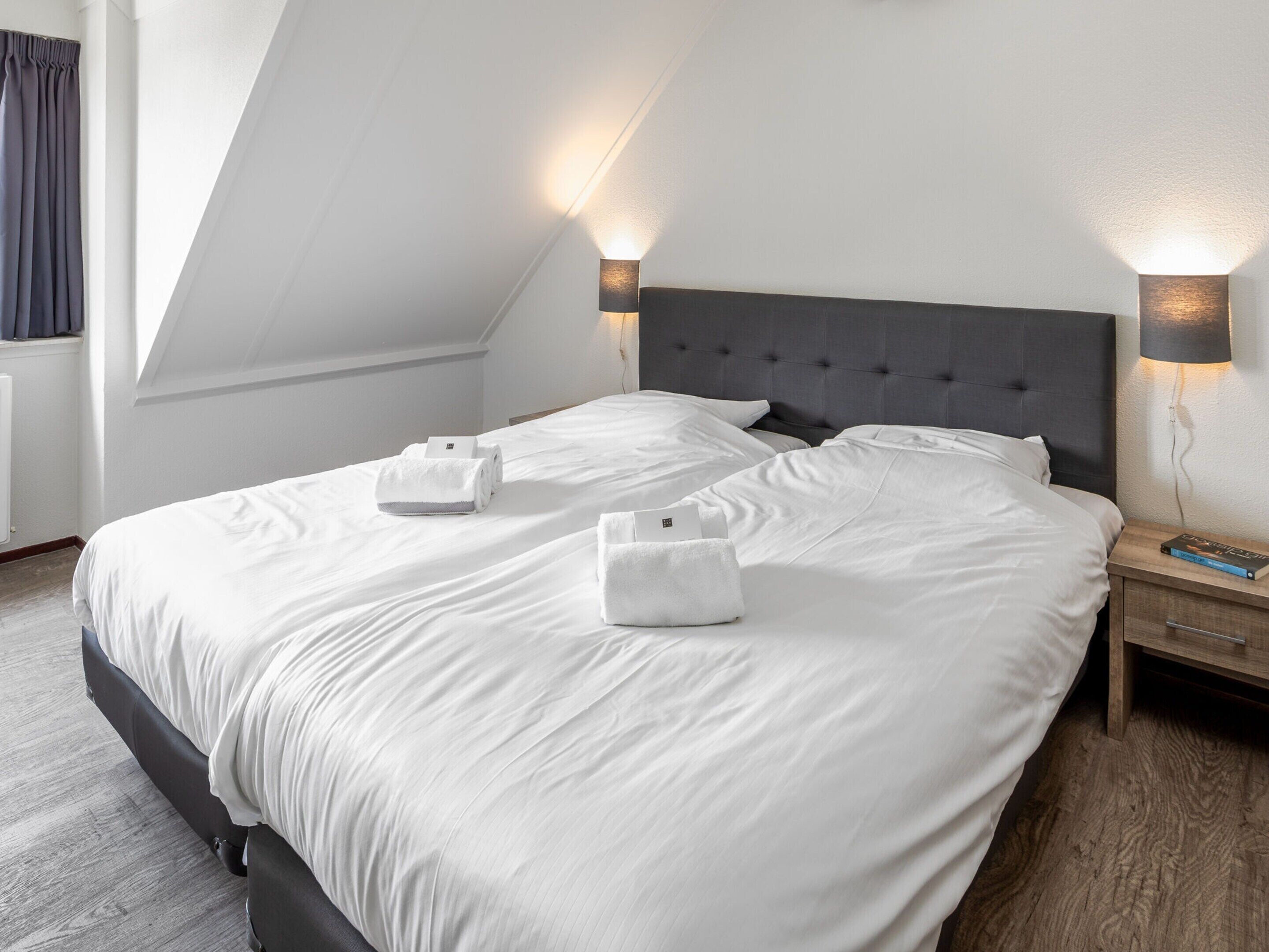 A neatly made bed with white linens and two towels, a dark headboard, and two wall-mounted lamps on either side.