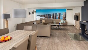 Een moderne woonkamer met een hoekbank, een eettafel met fruit, een salontafel en een groot schilderij van een kustlandschap.