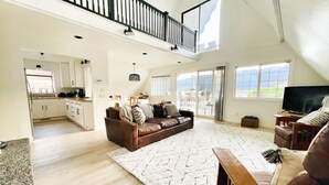 A spacious living area with a high ceiling, a brown leather sofa, a white rug, and a television on a wooden stand. - Lakefront, beach, hot tub, putt course, paddleboards, boat ramp, deck, fireplace (East Wenatchee)