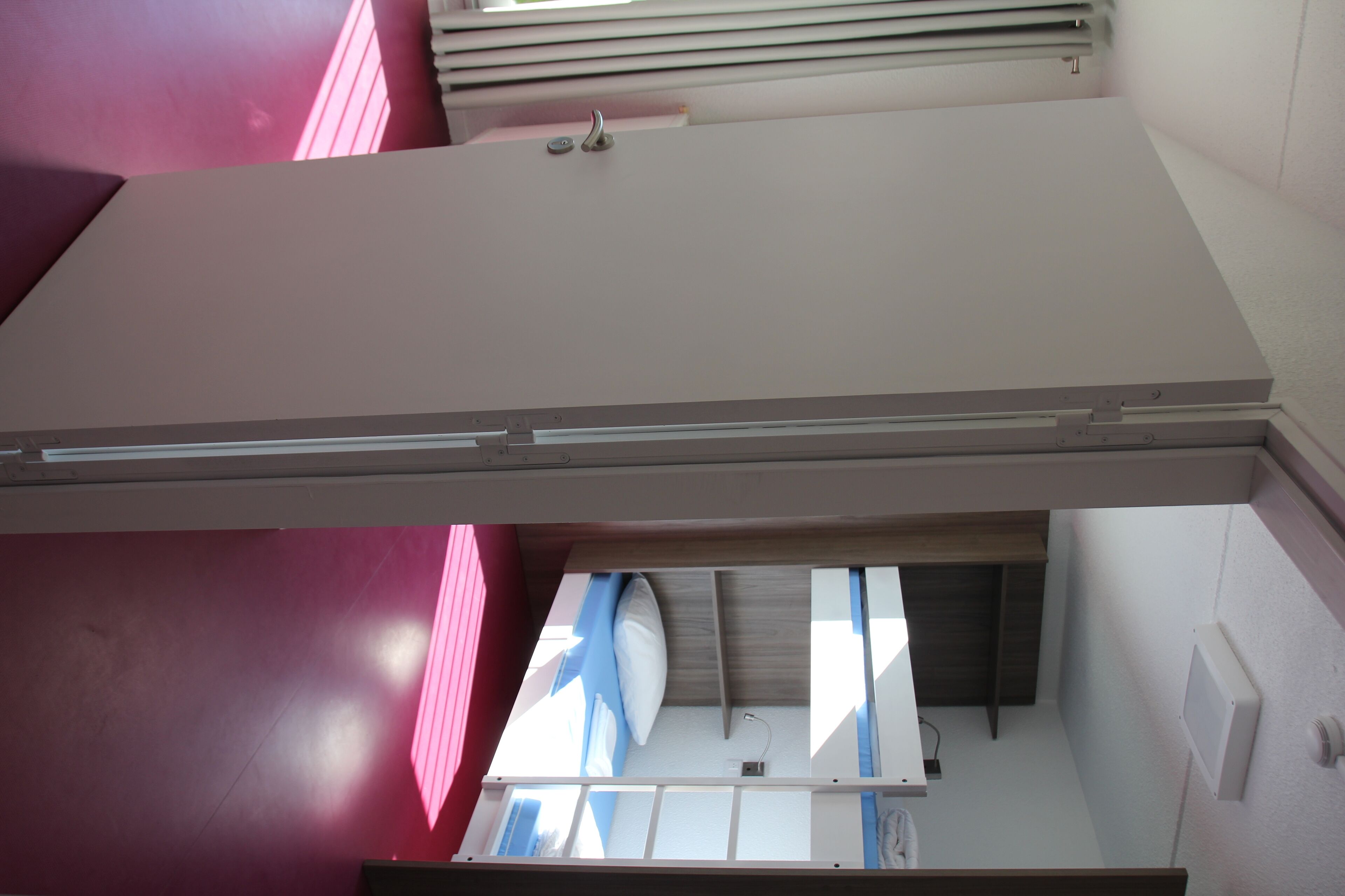 A room with a bed, a window with blinds, and a wall-mounted electrical outlet.