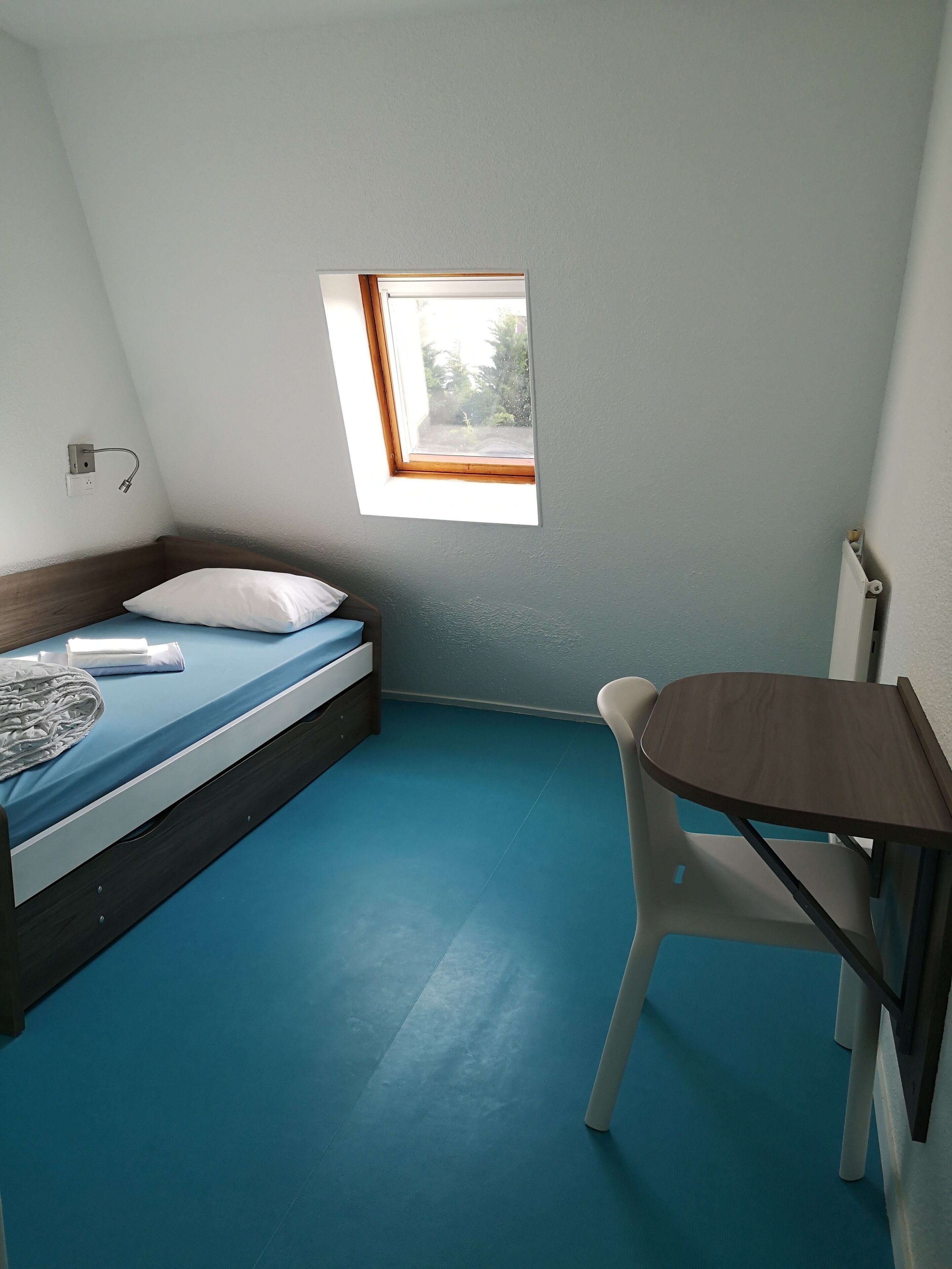 Una habitación pequeña y bien iluminada, con una cama individual, una mesita y una ventana que deja entrar luz natural.