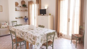A dining area with a table covered in a floral tablecloth, surrounded by chairs. There is a television on a cabinet, and a fan on the wall. - APP.TI VILLA ANNA - VILLA ANNA - TRILO PRIMO PIANO (Capoliveri)