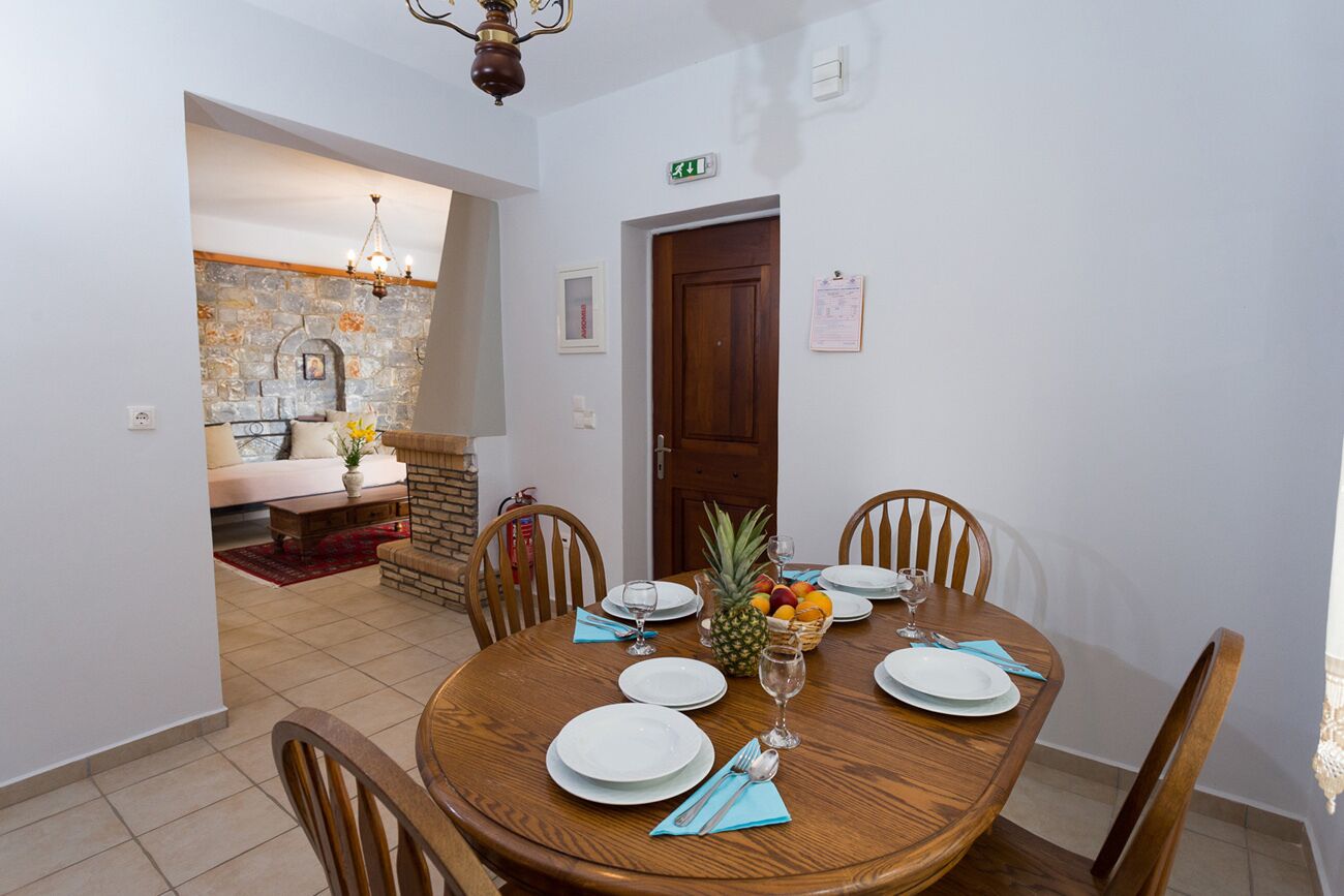 A dining area with a round wooden table set for four, surrounded by wooden chairs. A stone wall and a wooden door are visible in the background.