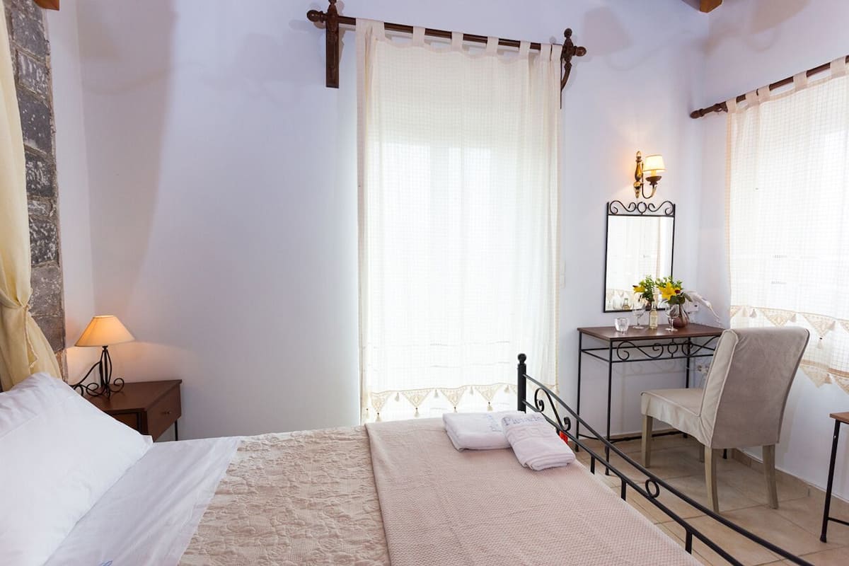 A bedroom with a bed, a nightstand, a desk with a chair, a mirror, and a window with curtains.