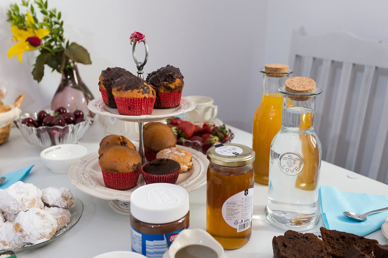 Et bord med forskellige bagværk, blandt andet muffins og cupcakes, et honningglas, en vandflaske og et glas appelsinjuice.