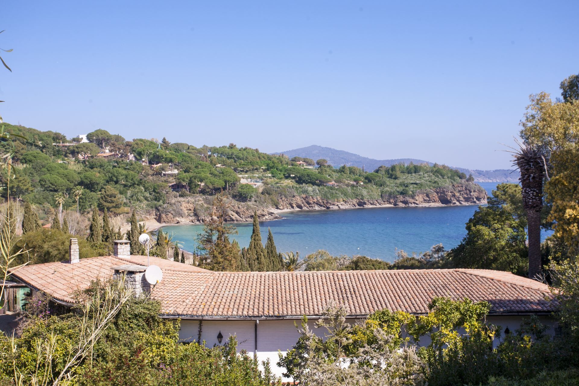 Una propiedad costera con tejado de baldosas, rodeada de vegetación y con vista a una bahía con una montaña al fondo.