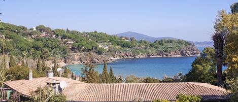 A coastal property with a tiled roof, surrounded by greenery and overlooking a bay with a mountain in the background.
