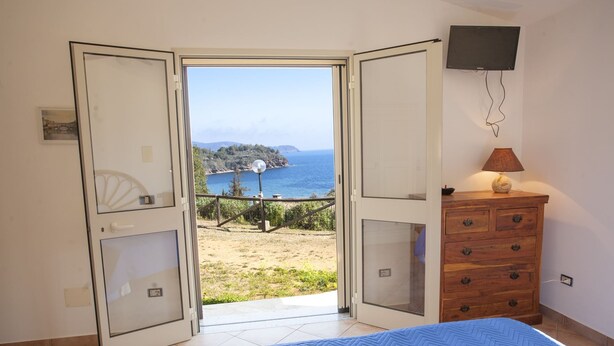 Een slaapkamer met een bed, een houten dressoir, een aan de muur bevestigde televisie en een balkon met uitzicht op zee.