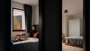A bedroom with a bed, a bedside table with a lamp, a window with curtains, and a view of a forest.