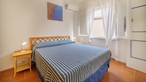 A bedroom with a bed, a nightstand, a wall-mounted air conditioner, a window with white curtains, and a door with a blue stripe. - APP.TI BAIA BLU - BAIA BLU 3 (Portoferraio)