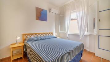 A bedroom with a bed, a nightstand, a wall-mounted air conditioner, a window with white curtains, and a door with a blue stripe.
