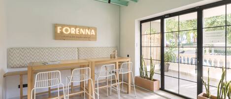Una habitación con una mesa de madera y cuatro sillas blancas, un letrero que dice FORENNA, y ventanas amplias con vistas al exterior.