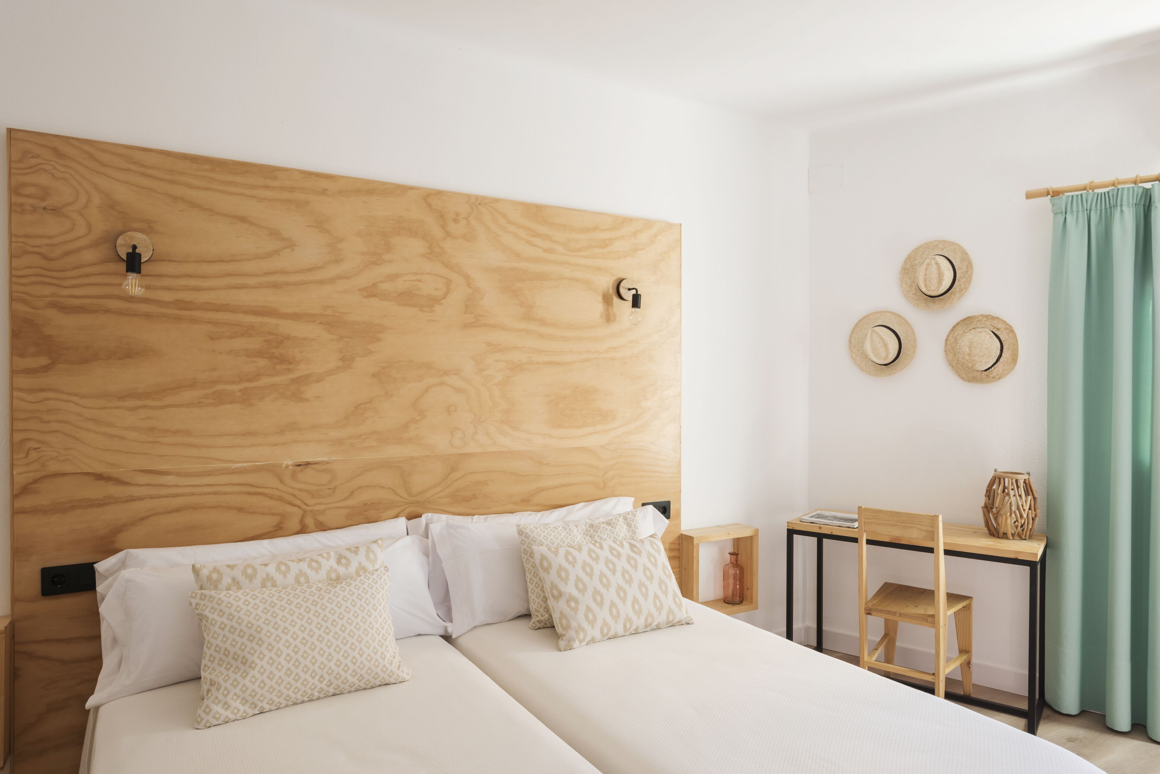 A bedroom with a wooden headboard, two beds with patterned pillows, a small table with a chair, and three hats on the wall.