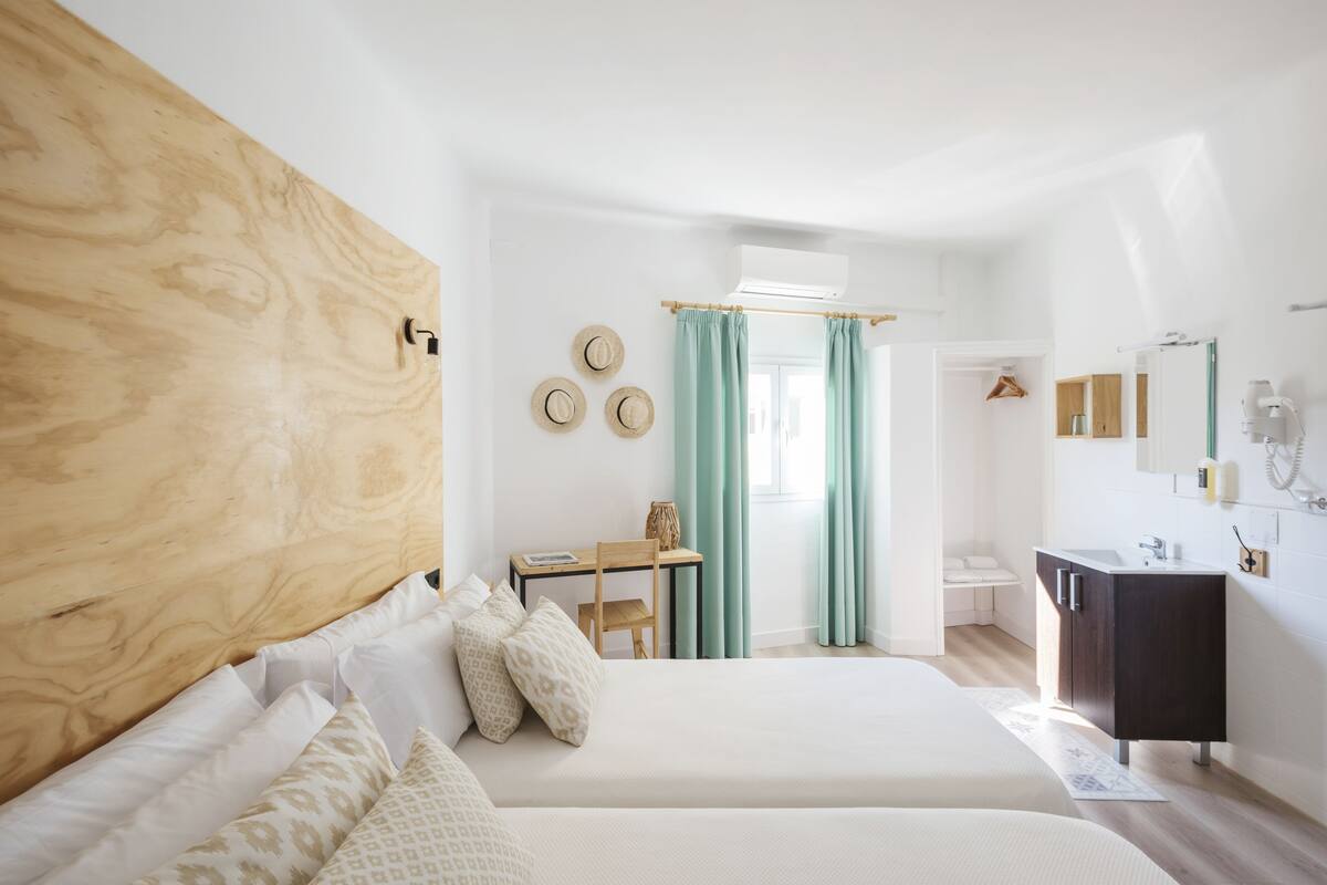 Ein modernes Schlafzimmer mit zwei Betten, einem Holzkopfteil, einem Schreibtisch mit Stuhl, einem Schminktisch mit Spiegel und einem Fenster mit Vorhängen.