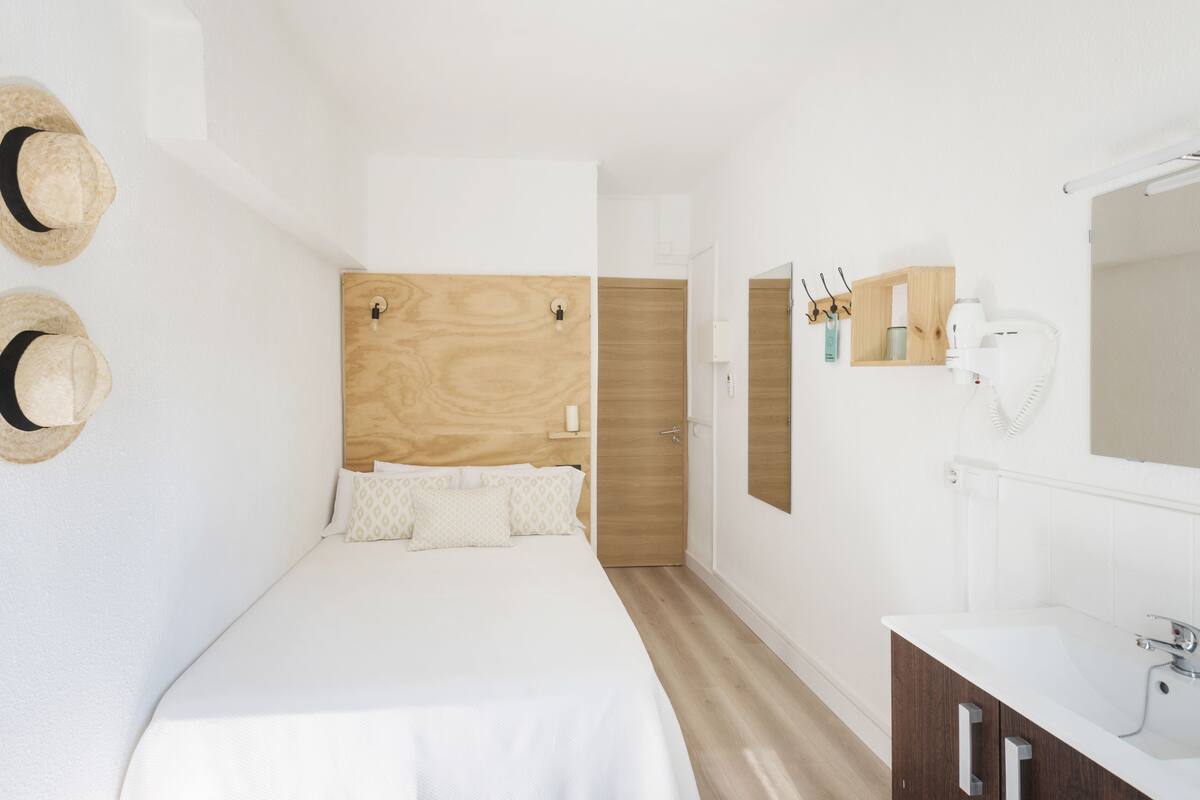 Ein Schlafzimmer mit einem Holz-Kopfteil, einem weißen Bett, Holzboden, einem weißen Badezimmer mit Spiegel und zwei Hüten an der Wand.