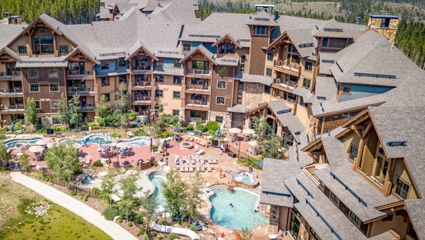 Un resort con varias edificaciones, un área de piscina central con tumbonas y un telón de fondo montañoso.