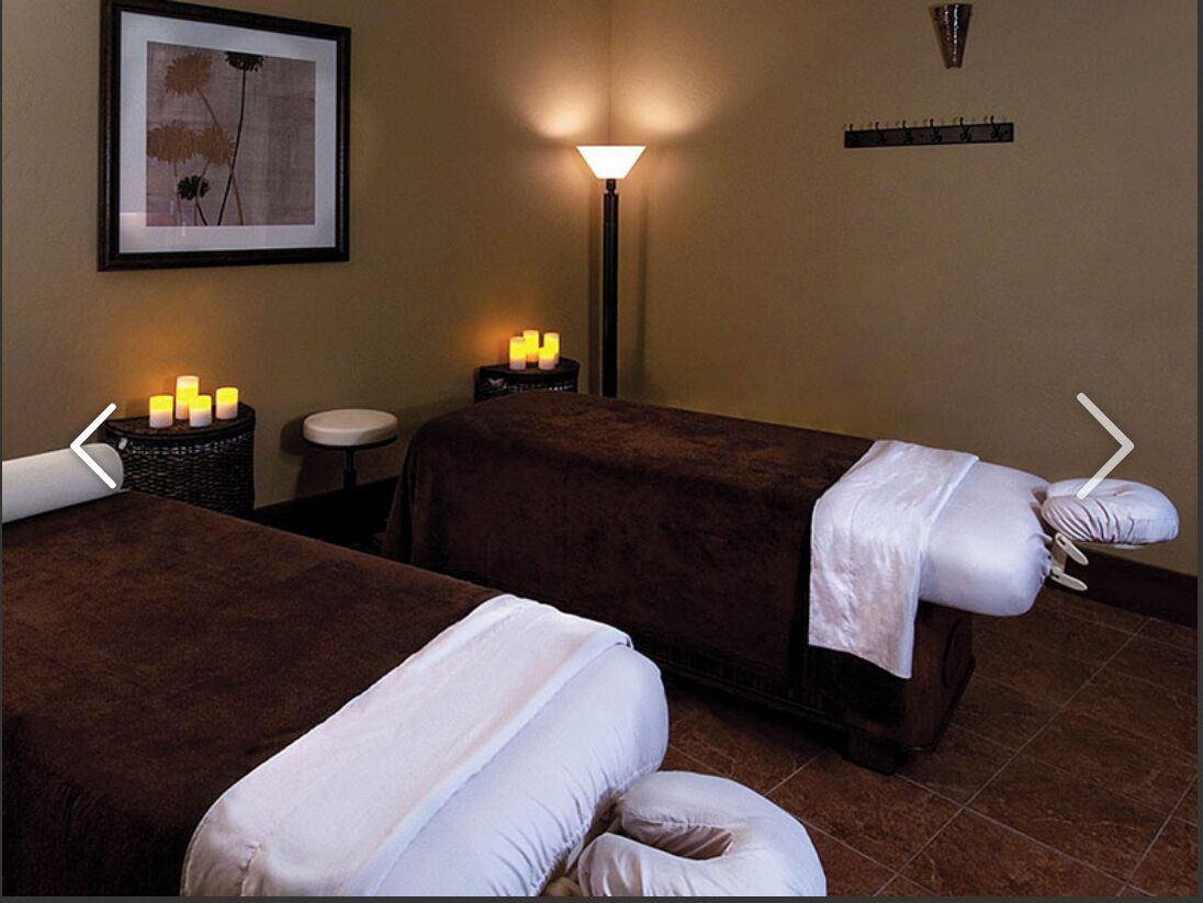 Ein Spa-Raum mit zwei Massagetischen, einem Wandbild, einer Stehlampe und einem kleinen Tisch mit Kerzen.