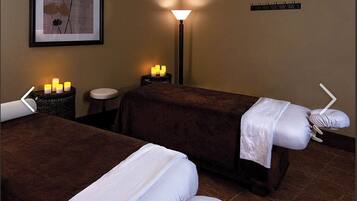 Ein Spa-Raum mit zwei Massagetischen, einem Wandbild, einer Stehlampe und einem kleinen Tisch mit Kerzen.