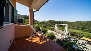 A balcony with two white lounge chairs, a small table, and a view of a vineyard and hills. - APP.TI CASA SERENA - CASA SERENA BILO 3 (Capoliveri)