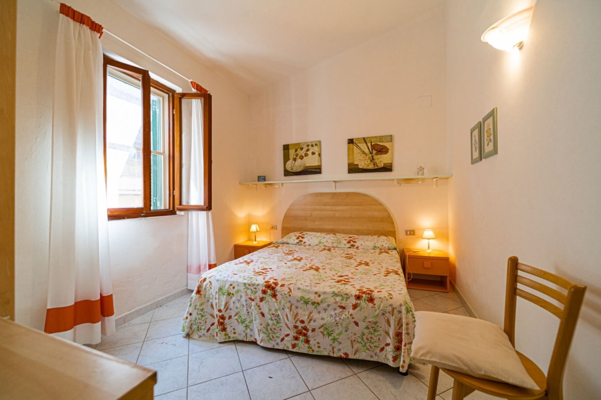 Une chambre à coucher comprenant un lit, deux tables de chevet, une chaise, une fenêtre avec des rideaux et des lampes fixées au mur.