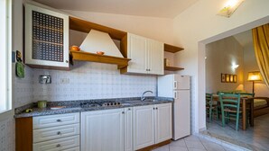A kitchen with white cabinets, a granite countertop, a stove, and a refrigerator. There is a dining area with a table and chairs visible through an open doorway. - APP.TI CASA SERENA - CASA SERENA TRILO 4 (Capoliveri)