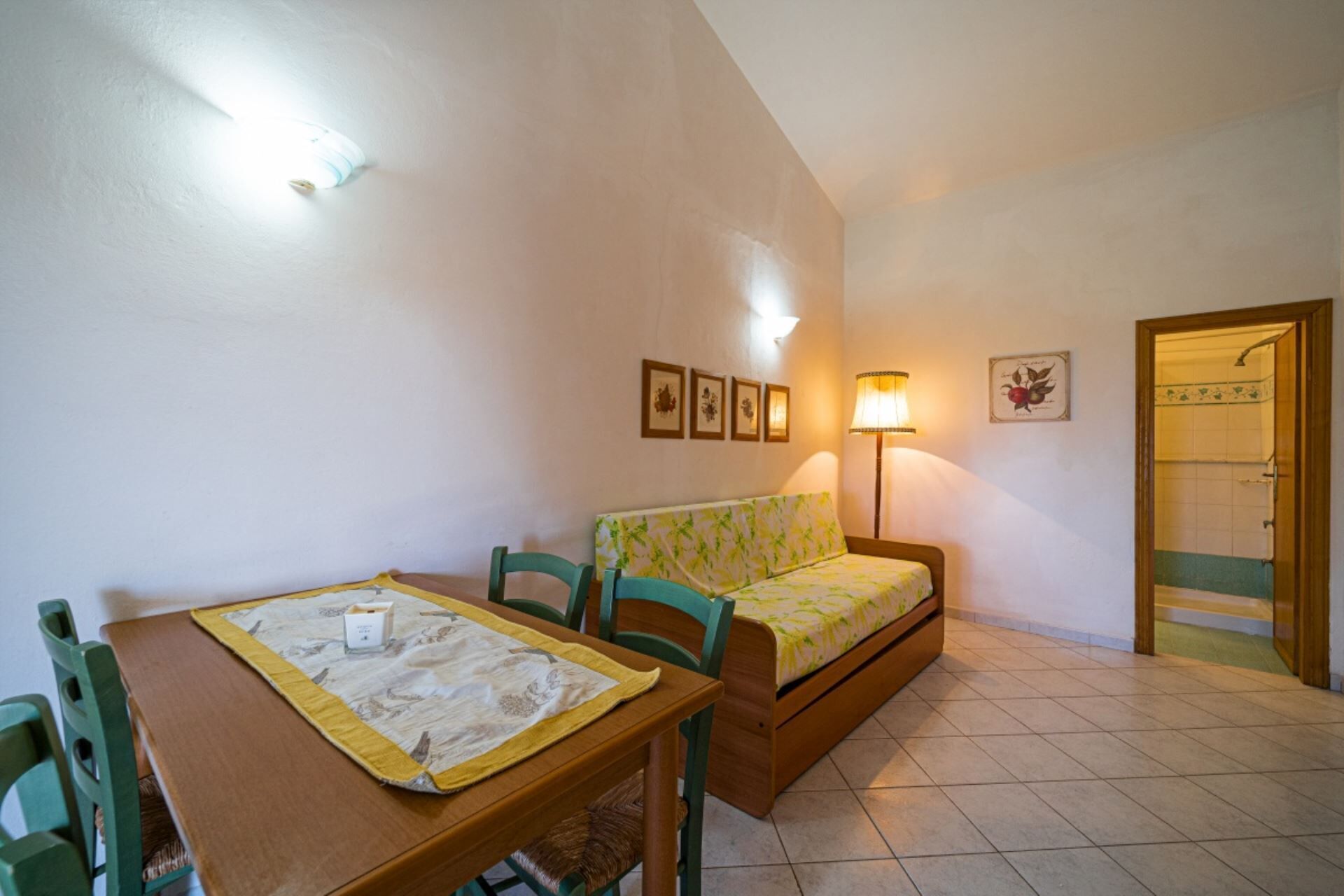 A small, cozy room with a wooden table, two chairs, a sofa, and a bed. There are framed pictures on the wall, a lamp, and a door leading to a bathroom.