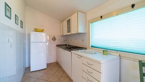 Une cuisine de taille réduite avec des meubles blancs, un réfrigérateur blanc, une cuisinière à gaz et une fenêtre équipée de stores.