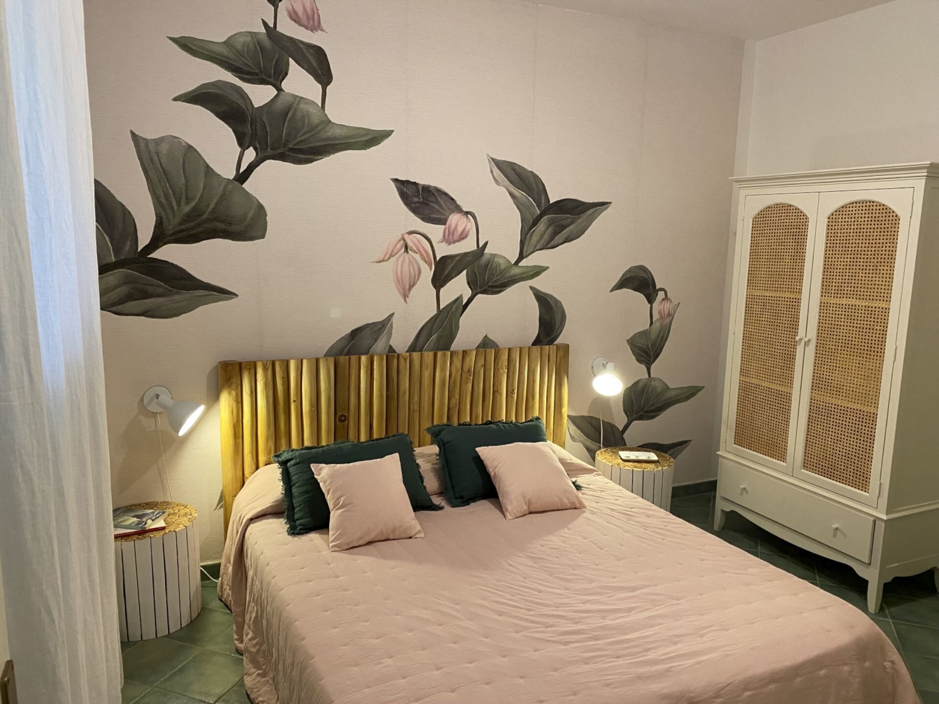 A bedroom with a bed, a headboard, a wardrobe, and a wall with a floral mural.