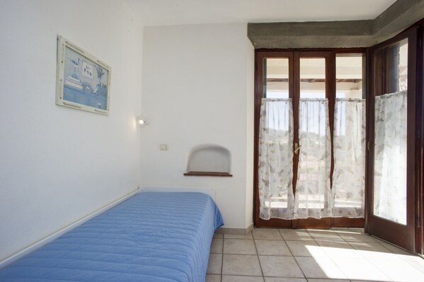 Una habitación con una cama, un pequeño estante, una ventana con cortinas y una puerta.