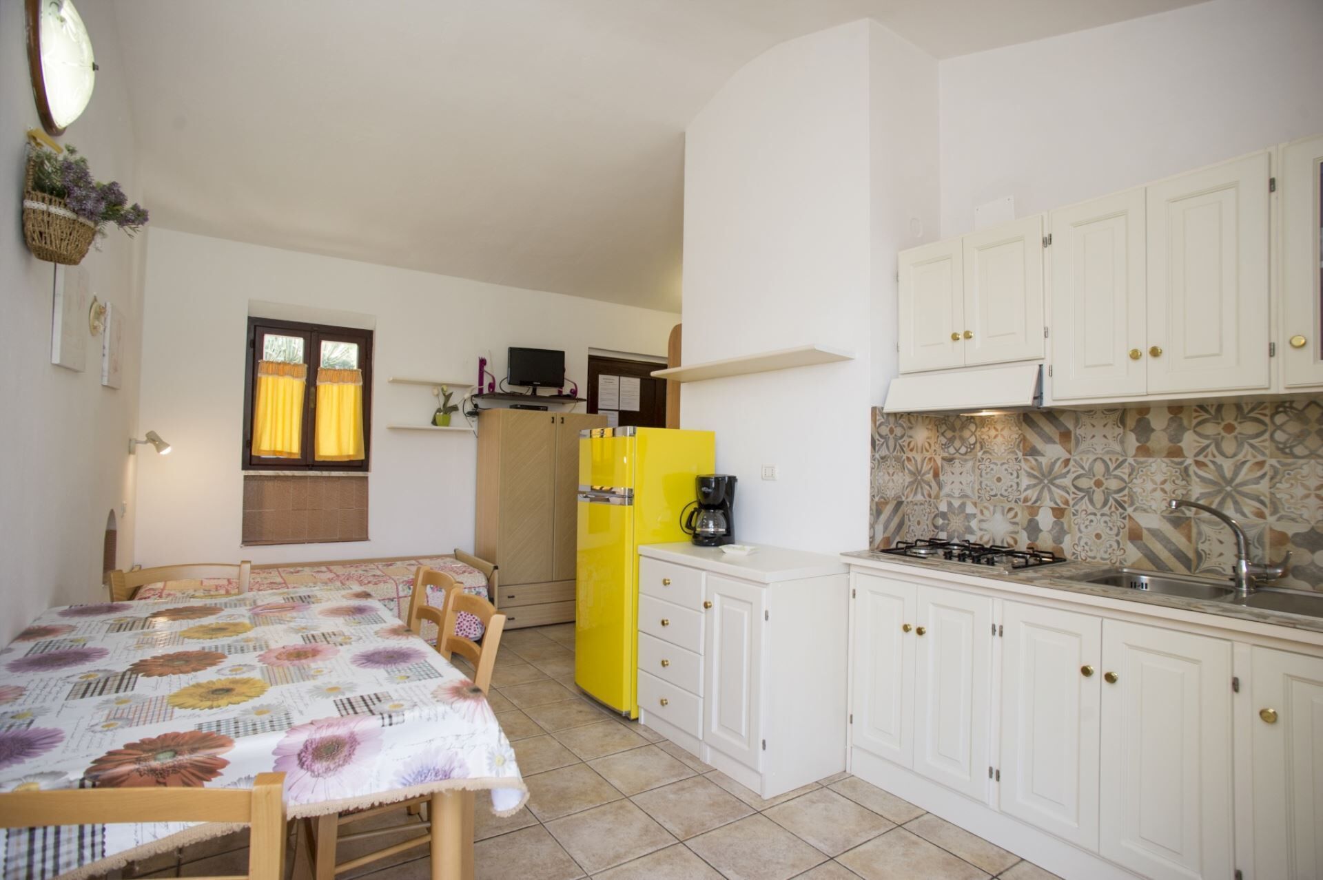 Une cuisine compacte avec des meubles blancs, un réfrigérateur jaune, une cuisinière à gaz et un coin repas agrémenté d’une nappe à motifs floraux.