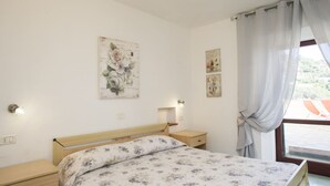 A bedroom with a wooden bed, a nightstand, a wall-mounted lamp, and a window with curtains. - APP.TI MORCONE - MORCONE BILO 1 (Capoliveri)