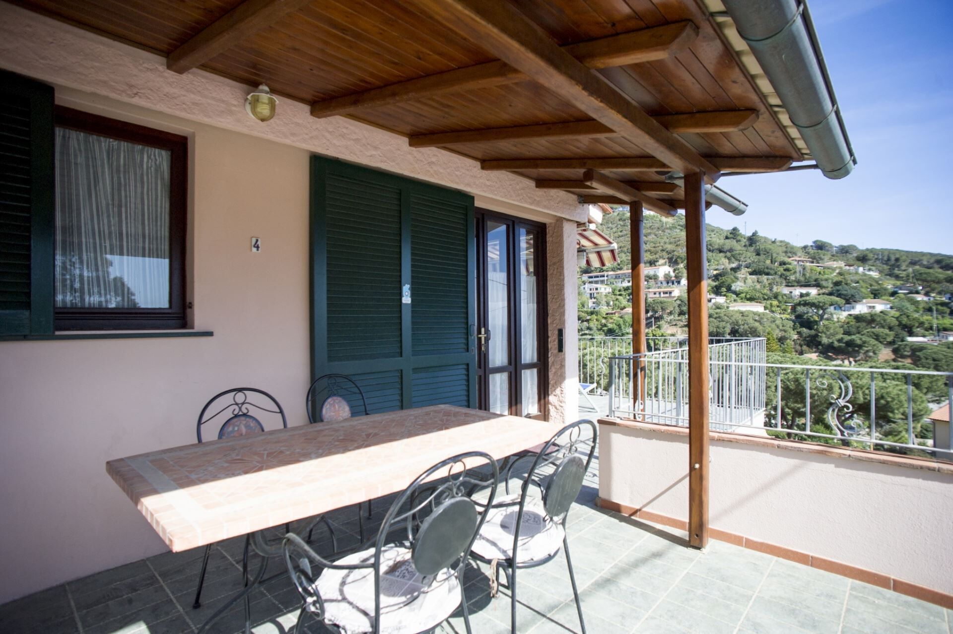 A balcony with a table and chairs, a view of a hillside, and a building with shutters.