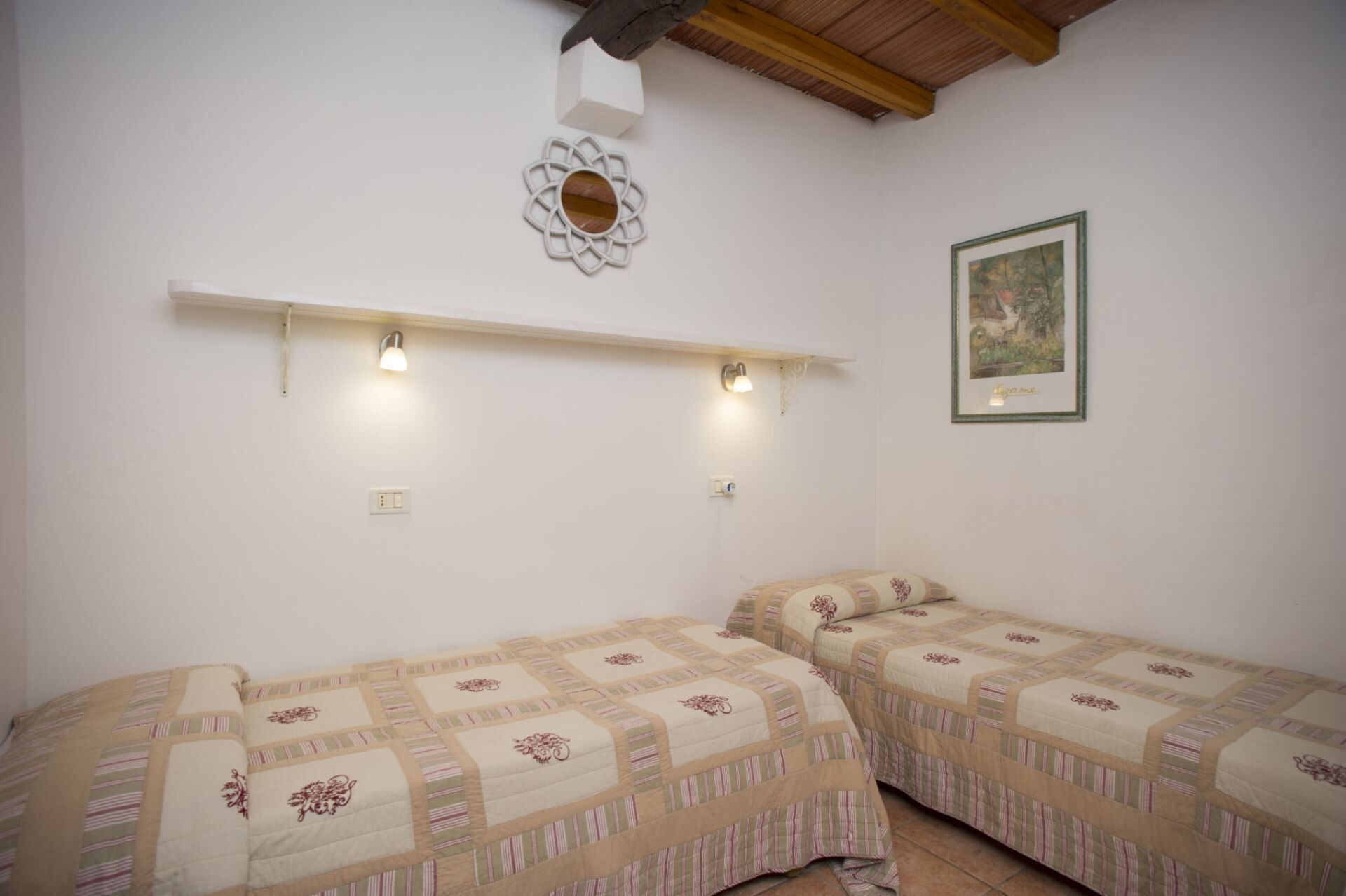 A room with two beds, each with a patterned bedspread, a wall-mounted shelf with lights, and a framed picture on the wall.