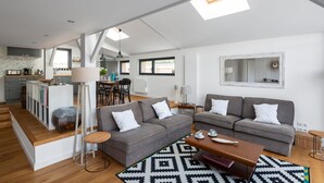 A modern apartment with a kitchen, dining area, and living room. It features a loft-style ceiling with skylights, grey sofas, and a patterned rug. - Villa Emeraude, Sublime maison à St Briac (Saint-Briac-sur-Mer)