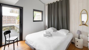 A bedroom with a bed, a chair, a nightstand, a window, and a curtain. - Villa Emeraude, Sublime maison à St Briac (Saint-Briac-sur-Mer)