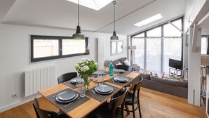 A modern dining area with a wooden table set for four, black chairs, and a large window with blinds. - Villa Emeraude, Sublime maison à St Briac (Saint-Briac-sur-Mer)
