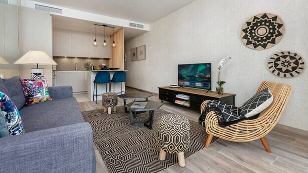 Un salon moderne avec un canapé gris, un fauteuil en osier, un téléviseur et une cuisine en arrière-plan.