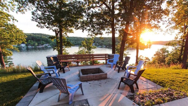 An outdoor seating area with a bench and chairs, a fire pit, and a view of a lake with boats. - A 5500 sq ft.Luxury Home~Huge Boat Dock~Perfect for Large Groups~Waterfront~Pool Table~Firepit (Camdenton)