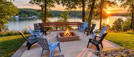 An outdoor seating area with a fire pit, surrounded by Adirondack chairs, overlooking a lake with a sunset in the background.