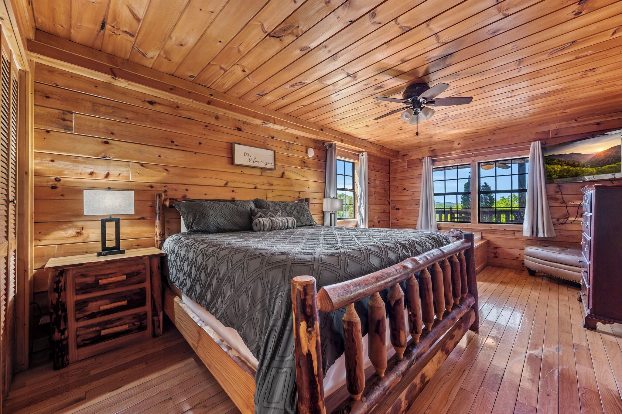 A wooden cabin bedroom with a bed, bedside table, lamp, and a view of the outdoors through large windows.