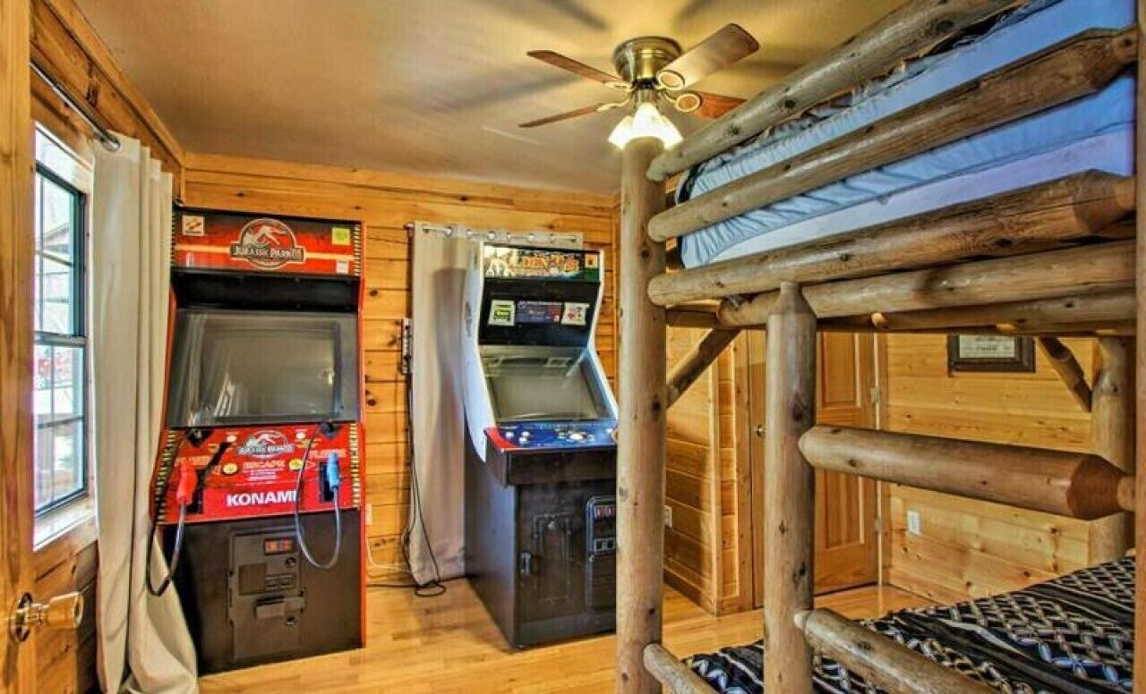 A wooden cabin interior with bunk beds, a ceiling fan, and arcade machines.