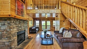 A spacious log cabin interior with a stone fireplace, a large leather sofa, and a staircase leading to an upper level. - Mountain View from front Porch and private Hot tub on the back porch! (Sevierville)