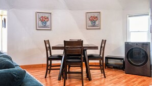 Een eetgedeelte met een houten tafel en stoelen, twee lijstwerken met bloemstillevens aan de muur, een wasmachine en een raam.