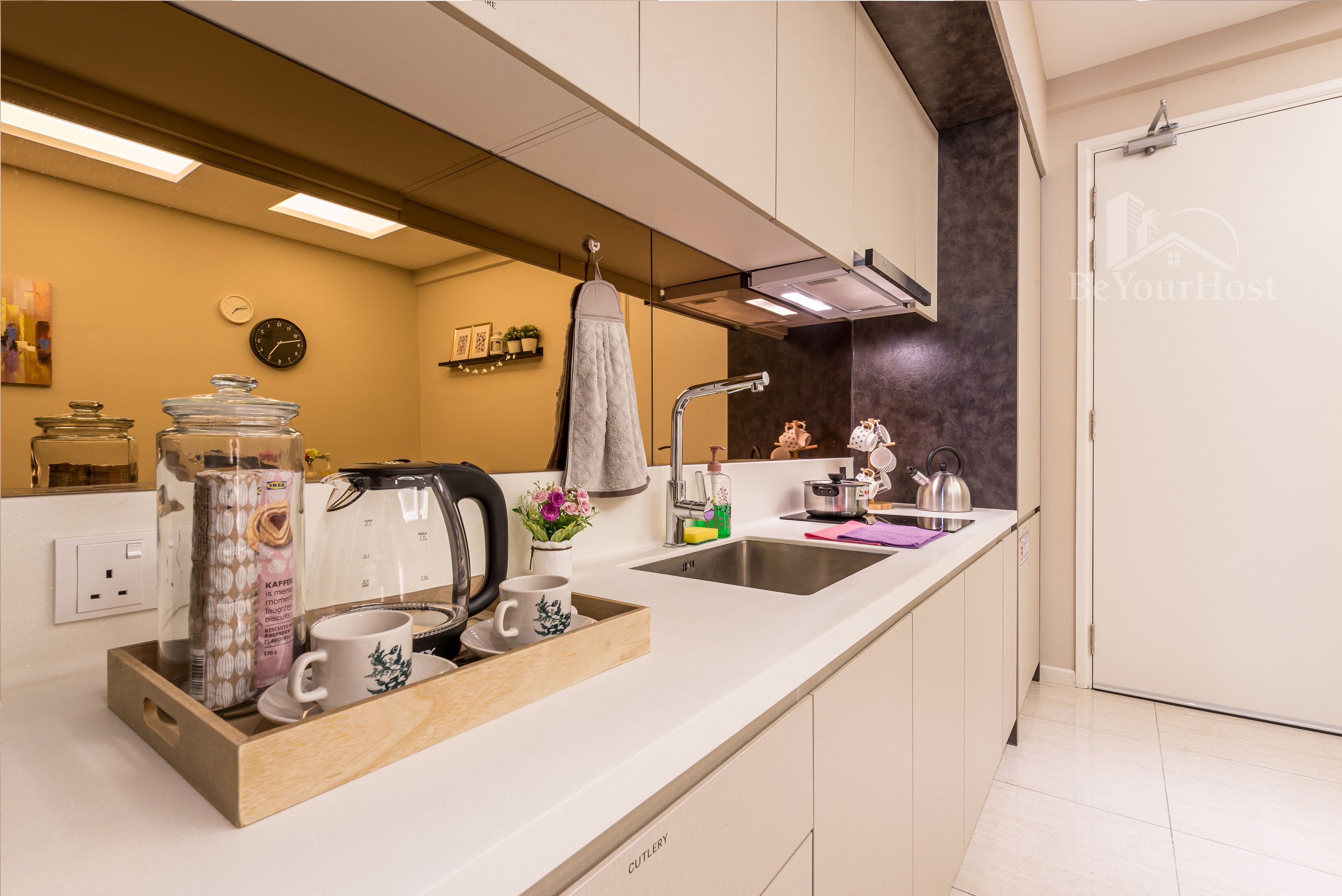 A modern kitchen with a white countertop, stainless steel sink, and a kettle. There are jars, a coffee maker, and a tray with cups and a teapot.