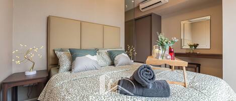 Una habitación de hotel moderna con una cama grande, una mesita de noche con lámpara, un espejo y un pequeño escritorio con un jarrón de flores.