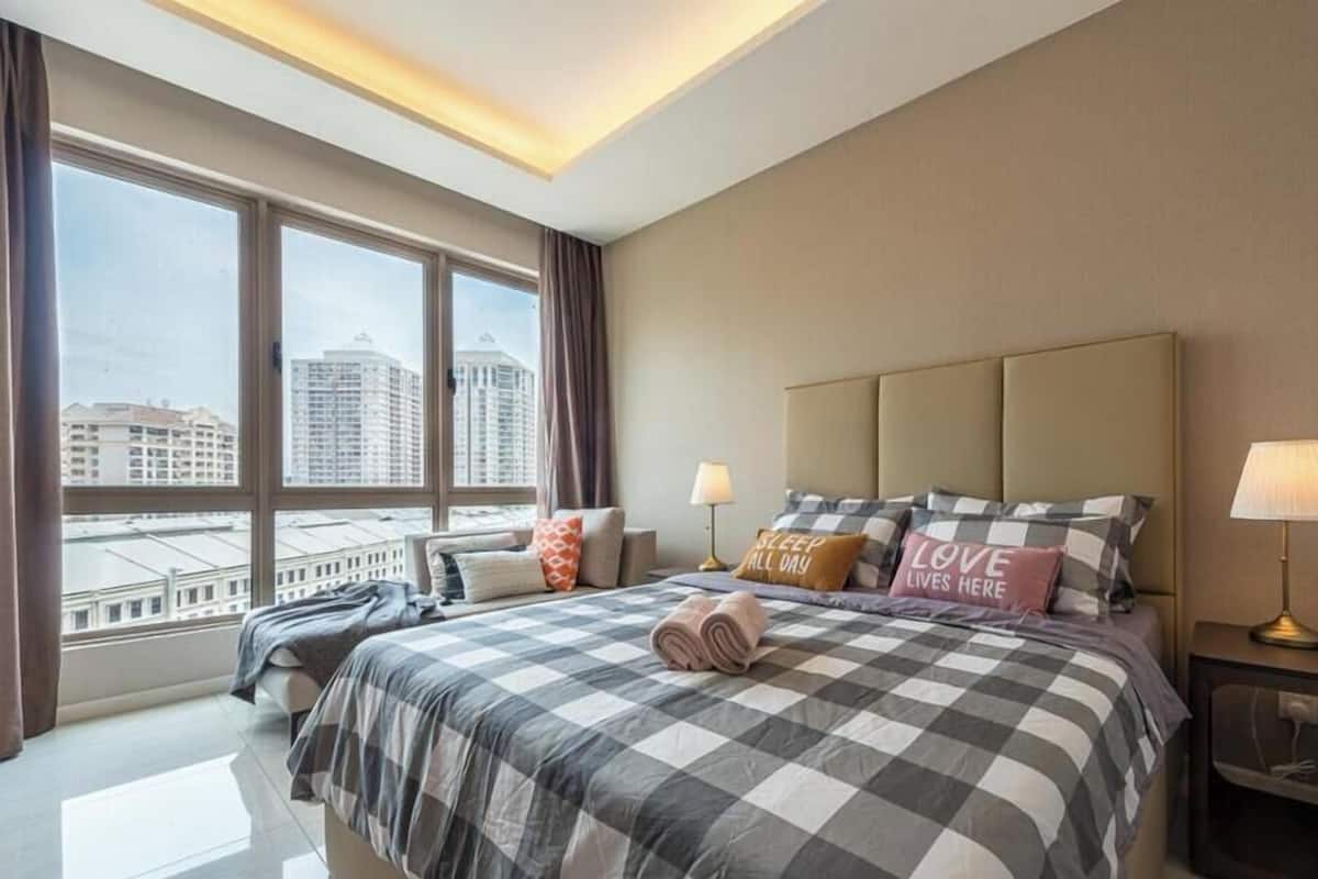 Ein Hotelzimmer mit einem großen Bett, zwei Nachttischlampen, Blick auf Gebäude durch große Fenster und einer Deckenleuchte.