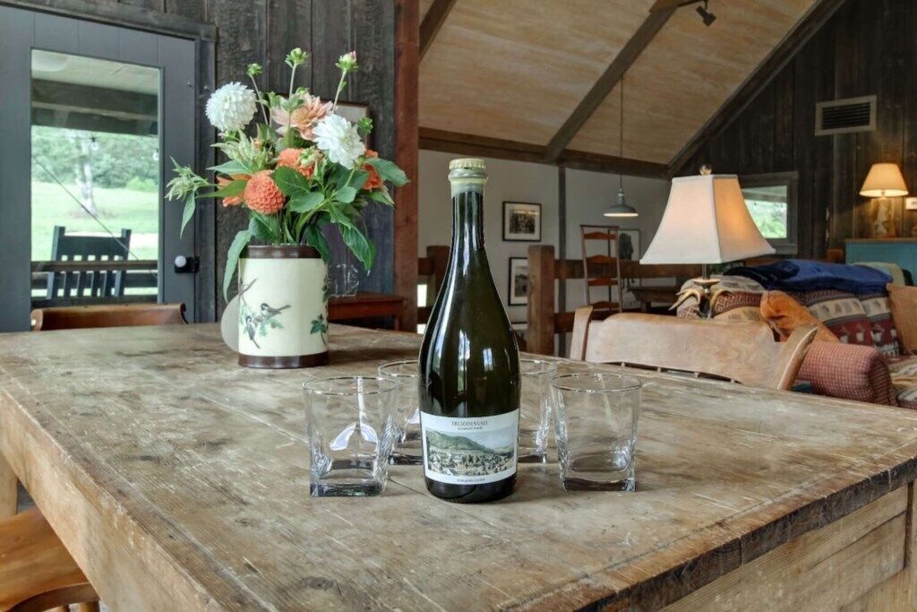 Ein rustikaler Holztisch mit einer Weinflasche, Gläsern und einer Blumenvase in ländlicher Umgebung.
