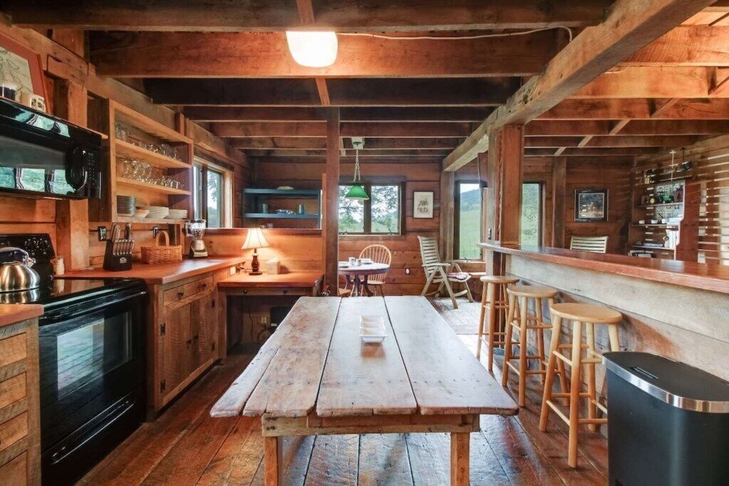 Ein rustikaler Holzinnenraum mit einem großen Holztisch, Barhocker, einer Küchenzeile mit Herd, Regalen mit Geschirr und einem Fenster, das natürliches Licht hereinlässt.