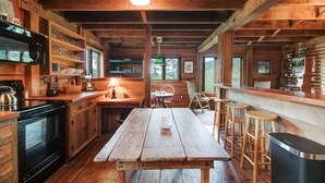 A rustic wooden interior with a large wooden table, bar stools, a kitchen area with a stove, shelves with dishes, and a window providing natural light.